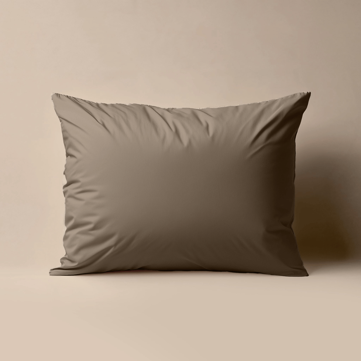 Een Zelesta Premiumbed Kussensloop in taupe, gemaakt van perkal katoen, staat rechtop tegen een lichtbeige achtergrond.