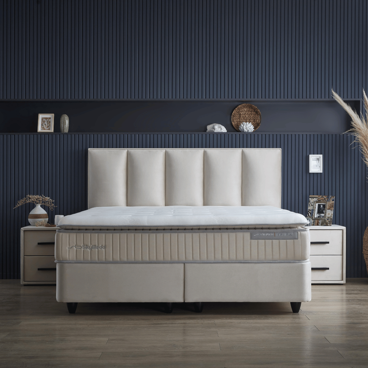 Een moderne slaapkamer heeft een luxe fluweelachtig beige gestoffeerd bed met een hoog hoofdeinde, geflankeerd door Zelesta SkyBedd Orion - Beige Nachtkastjes tegen een donkerblauwe accentmuur met panelen.