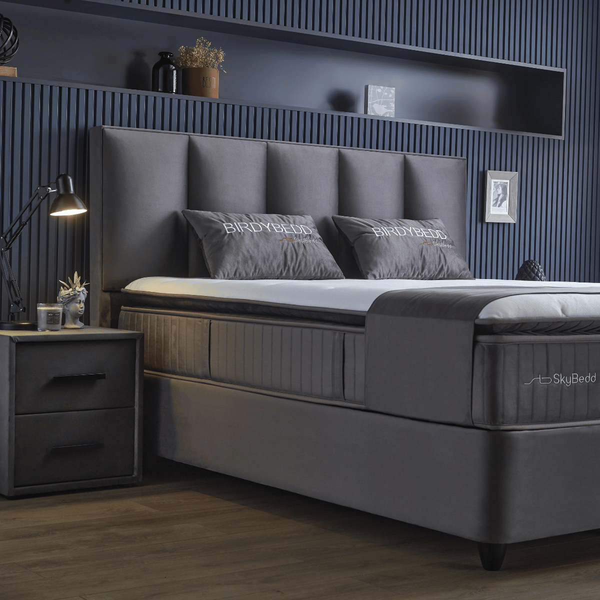 Moderne slaapkamer met een grijs gestoffeerd bed, bijpassend beddengoed en het Zelesta SkyBedd Orion - Antraciet nachtkastje naast een decoratieve plank op een donkerblauwe wand met panelen.