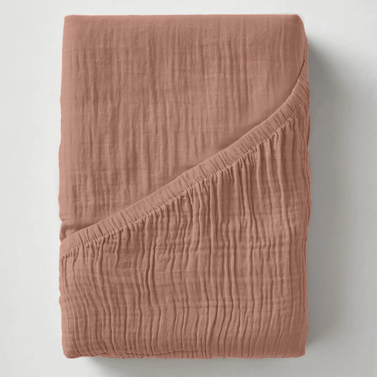 Het Mousseline Hoeslaken - Dusty-Rose, een rozekleurig hoeslaken van mousseline katoen ademend beddengoed, wordt getoond op een wit oppervlak met een hoek naar achteren gevouwen om de dikte en stofdetails te laten zien.