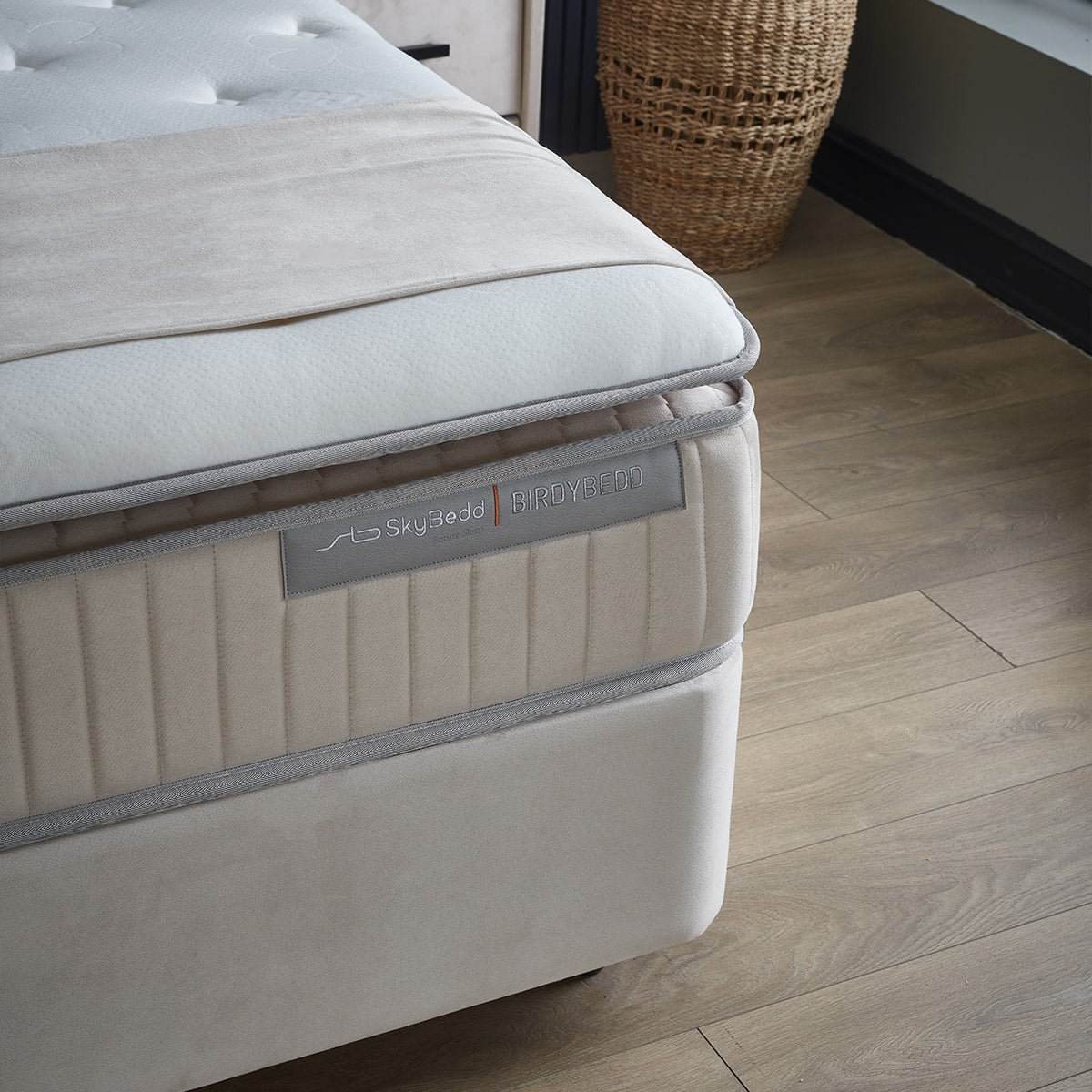 Een close-up van een beige Zelesta SkyBedd Orion matras met een gewatteerde bovenkant, geplaatst op een houten vloer naast een rieten mand.