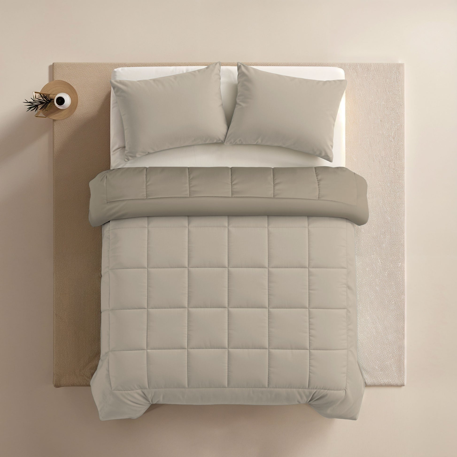 Bovenaanzicht van een netjes opgemaakt bed met het Zelesta Comfort Duo Dekbed in Taupe & Champagne, twee kussens en een klein rond nachtkastje met een enkele tak in een vaas.