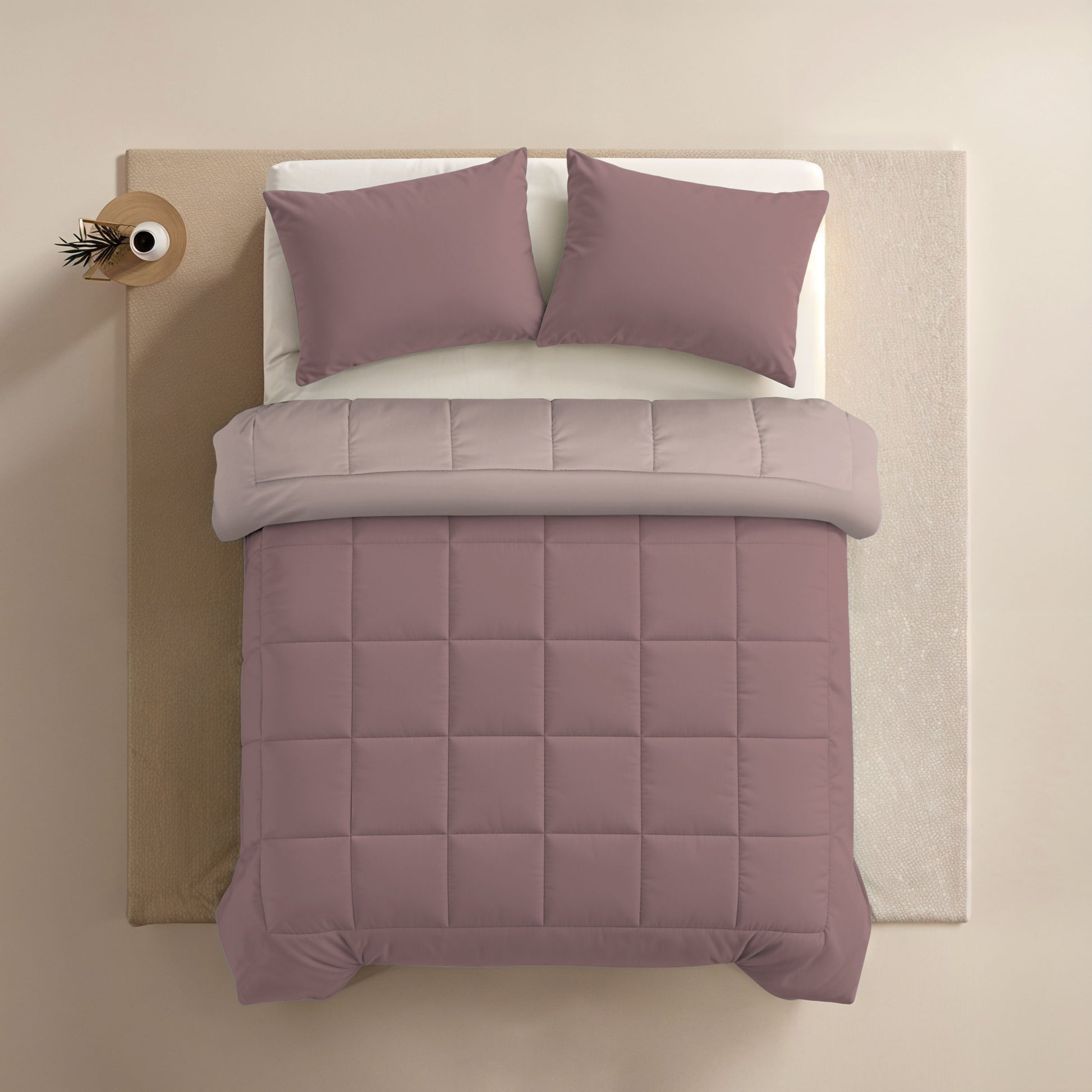 Netjes opgemaakt bed met mauve dekbed, twee kussens met Zelesta Comfort Duo Kussensloop - Oudroze (60x70cm), en een lichtgrijs bovenlaken, van bovenaf gezien. Een klein rond bijzettafeltje met een vaas staat naast het bed.
