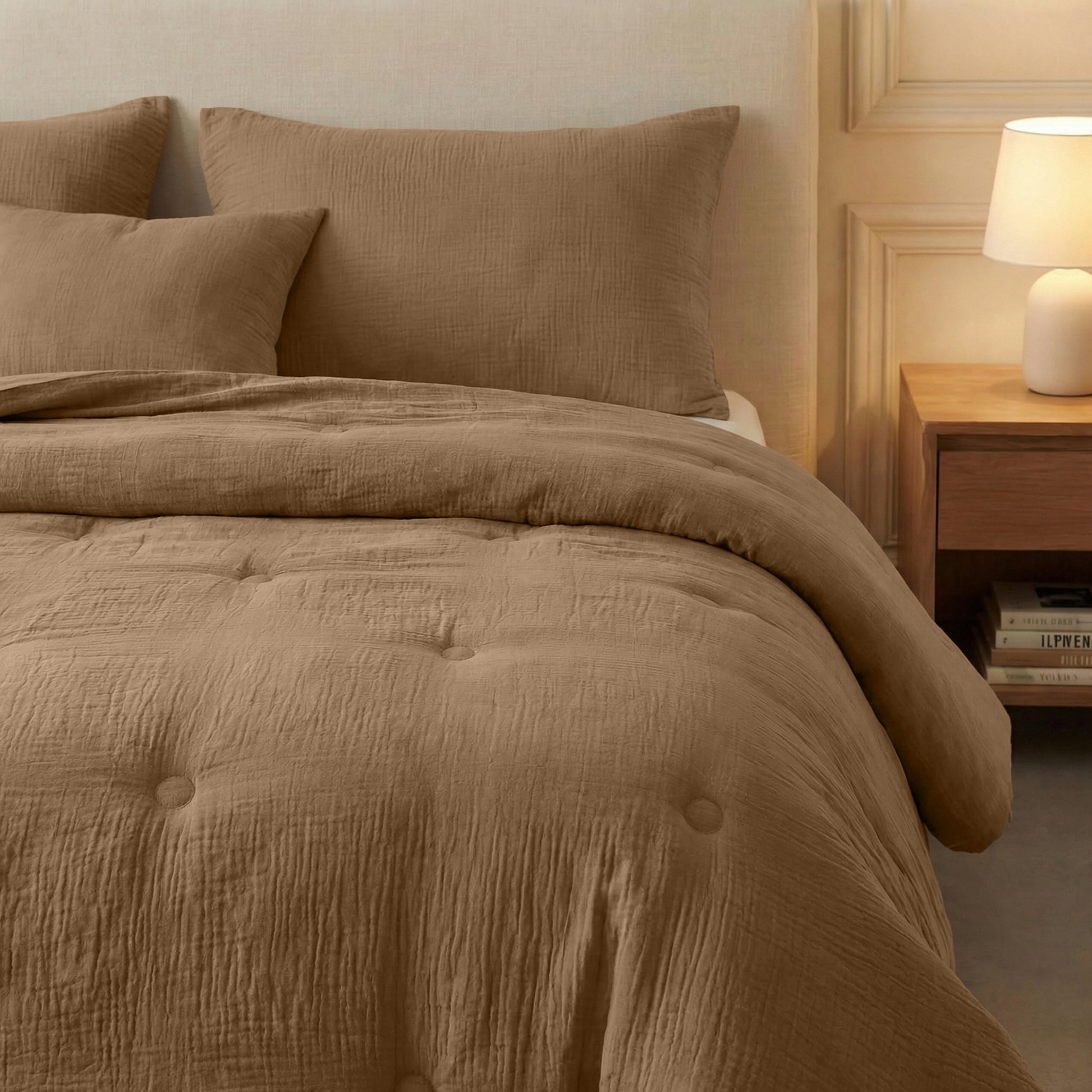 Een bed gekleed in het Zelesta Japandibed Katoen Mousseline - Bruin, met twee kussens en een Japandi dekbed, staat naast een houten nachtkastje met een lamp en gestapelde boeken in een zacht verlichte kamer.