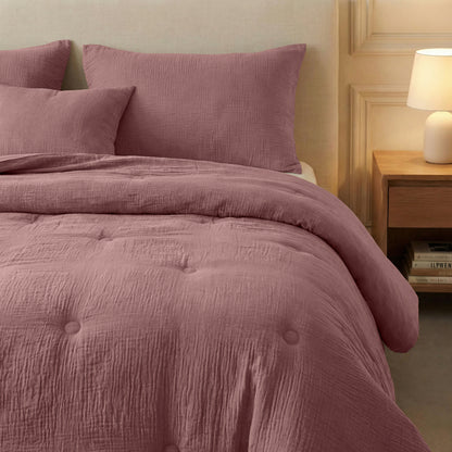 Een keurig opgemaakt bed met de Zelesta Japandibed Katoen Mousseline in Oudroze en kussens, naast een houten nachtkastje met een lamp en gestapelde boeken, tegen een beige muur.