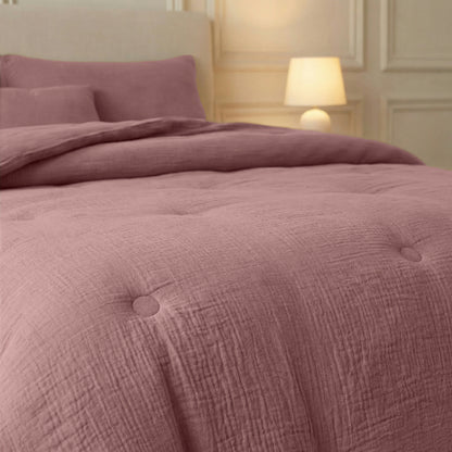 Een Zelesta Japandibed Katoen Mousseline - Oud Roze met bijpassende kussens staat in een zacht verlichte slaapkamer, met een lamp en een crèmekleurige wand op de achtergrond.