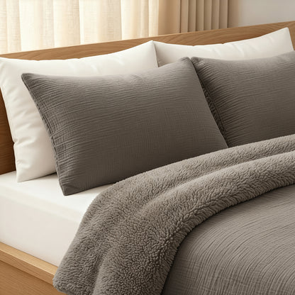 Een opgemaakt bed met witte kussens en lakens, twee grijze structuurkussens en het temperatuurregulerende Zelesta Teddy Katoen Mousseline - Taupe dekbed, in een kamer met lichte gordijnen.