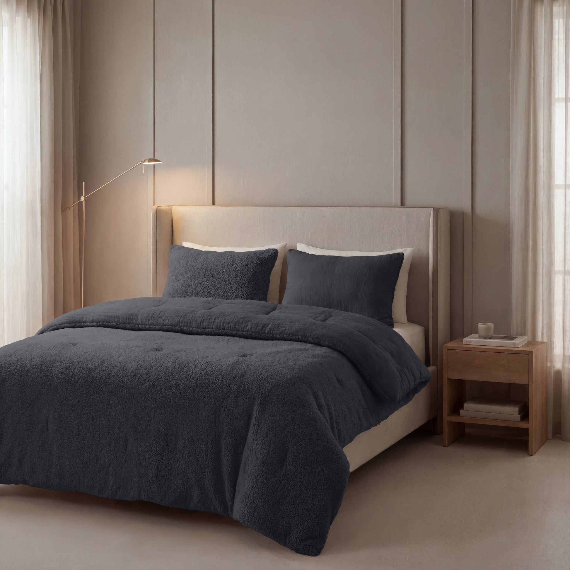 Een netjes opgemaakt bed met Zelesta Teddy Katoen Mousseline - Antraciet beddengoed en kussens staat tegen een lichtbeige muur, naast een houten nachtkastje en staande lamp in een zacht verlichte slaapkamer.