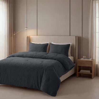 Een netjes opgemaakt bed met Zelesta Teddy Katoen Mousseline - Antraciet beddengoed en kussens staat tegen een lichtbeige muur, naast een houten nachtkastje en staande lamp in een zacht verlichte slaapkamer.