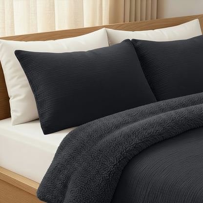 Een keurig opgemaakt bed met zwarte temperatuurregulerende kussens en het gezellige Zelesta Teddy Katoen Mousseline - Antraciet dekbed, tegen een houten hoofdeinde en lichtgekleurde gordijnen.