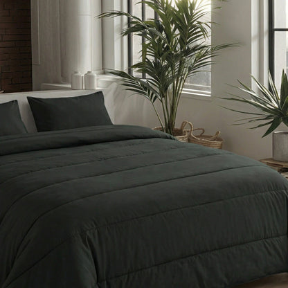Een netjes opgemaakt bed met het Zelesta Comfort Uni Dekbed - Zwart en kussens staat bij grote ramen en groene potplanten in een moderne, minimale slaapkamer - ideaal voor comfort het hele jaar door.