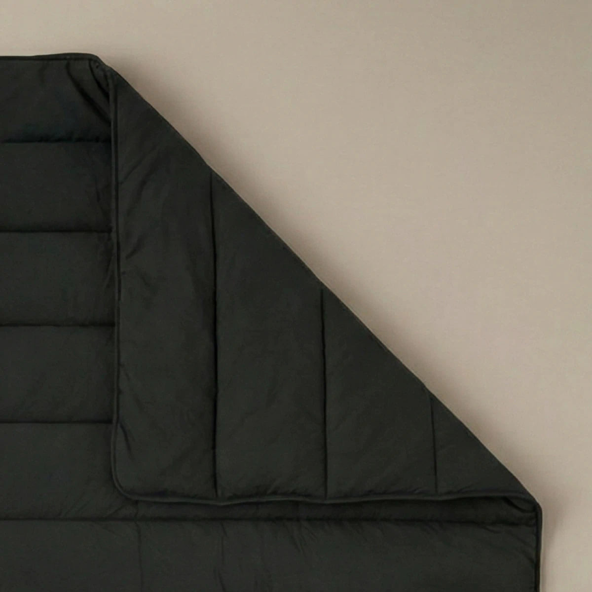 Het Zelesta Comfort Uni Dekbed - Zwart, een donkergrijze, doorgestikte all-season deken, is netjes uitgespreid op een beige ondergrond met één hoek omgevouwen om de binnenkant te laten zien.