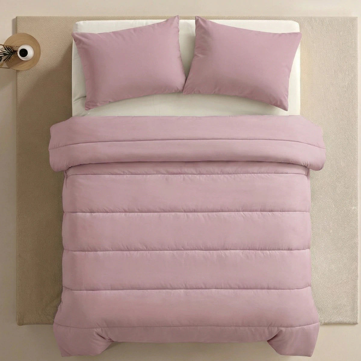 Een netjes opgemaakt bed met het Zelesta Comfort Uni Dekbed in Oud Roze en bijpassende kussens, op een crèmekleurig vloerkleed naast een ronde bijzettafel, benadrukt de tijdloze charme van klassieke dekbedden.