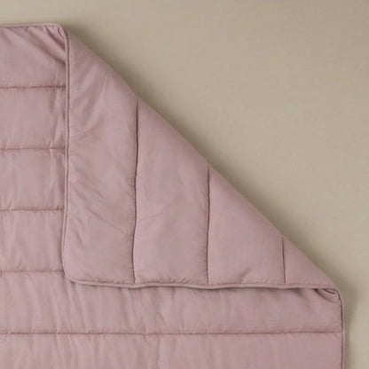 Het Zelesta Comfort Uni Dekbed in oud roze, een lichtroze doorgestikte all-season deken, wordt gedeeltelijk teruggevouwen getoond op een beige ondergrond.