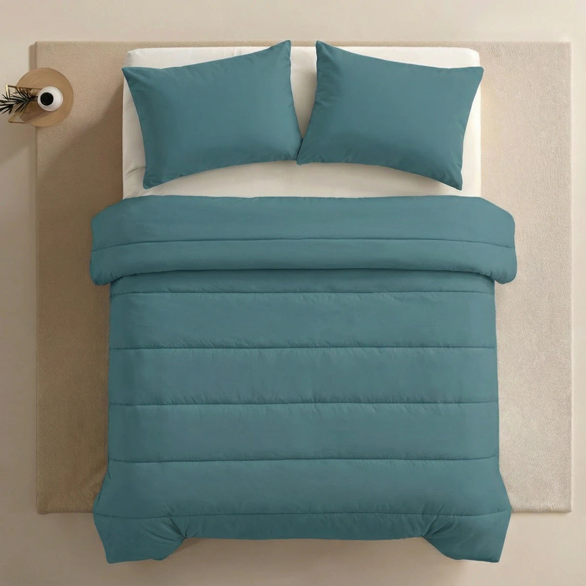 Een keurig opgemaakt bed met het Zelesta Comfort Uni Dekbed in zeegroen en twee bijpassende kussens, geplaatst op een crèmekleurig vloerkleed naast een klein rond nachtkastje met een vaas.