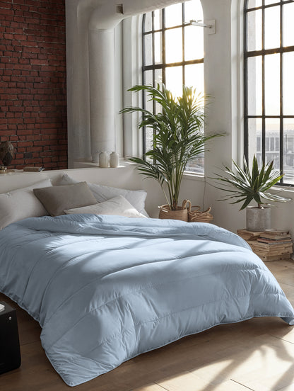 Een netjes opgemaakt bed met een Zelesta Everyday Uni Dekbed - Lichtblauw staat bij grote ramen in een zonovergoten kamer met bakstenen en witte muren, houten vloeren en potplanten.