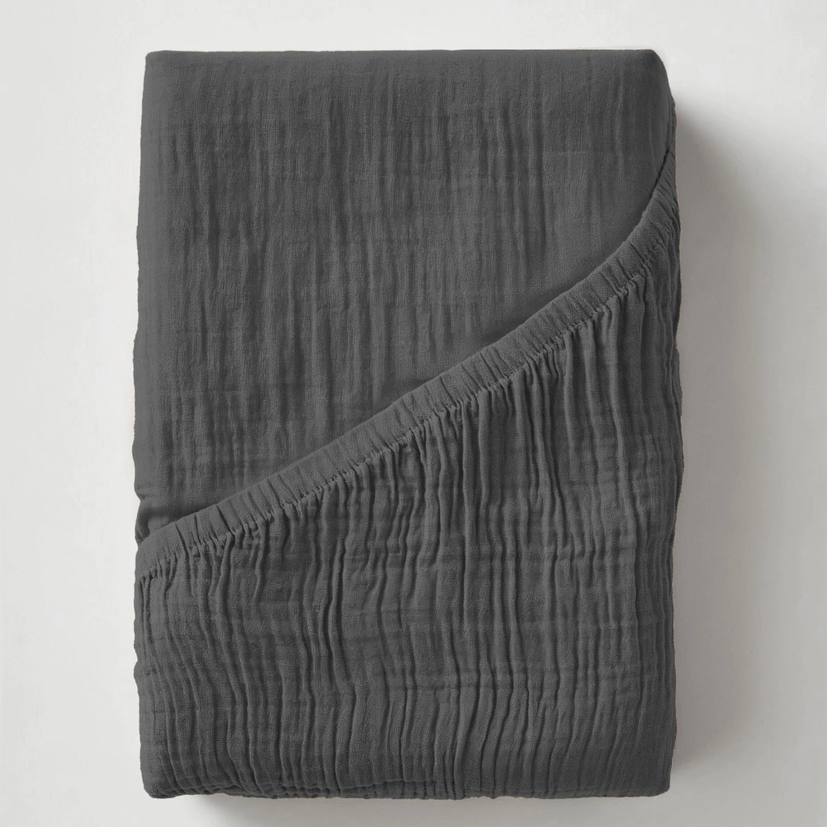 De Mousseline Hoeslaken - Antraciet is een donkergrijs, gekreukt lakentje van zacht mousseline katoen, netjes gevouwen met één hoek omgeslagen om het gestructureerde oppervlak te laten zien.
