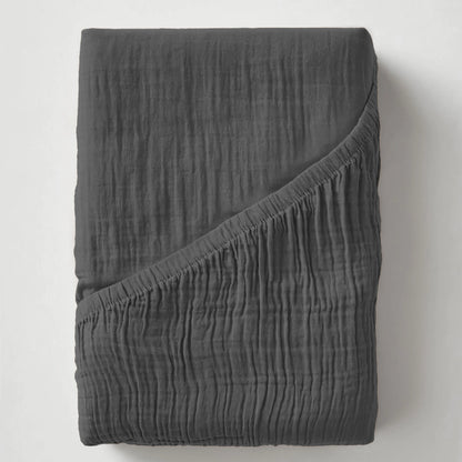 De Mousseline Hoeslaken - Antraciet is een donkergrijs, gekreukt lakentje van zacht mousseline katoen, netjes gevouwen met één hoek omgeslagen om het gestructureerde oppervlak te laten zien.