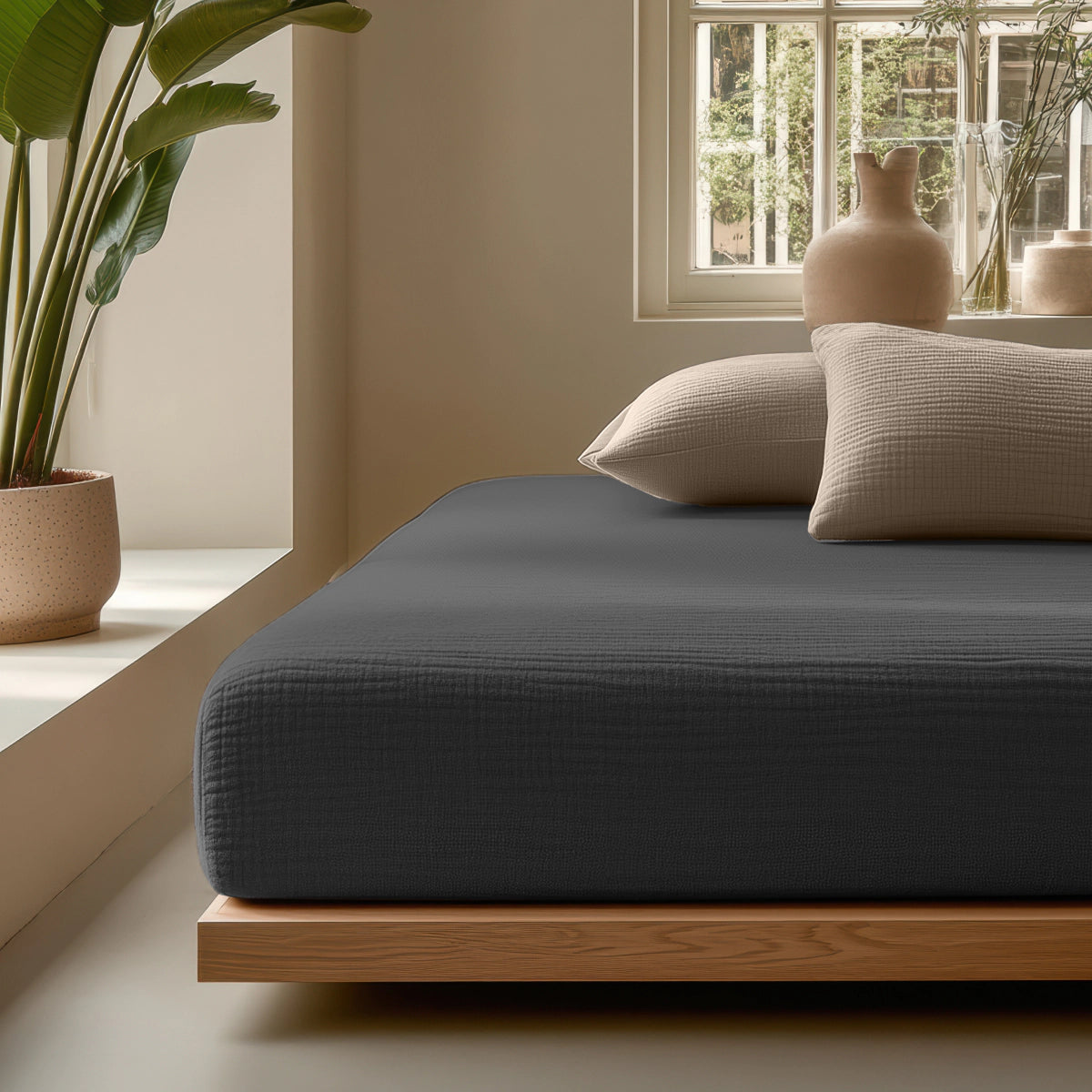 Een minimalistische slaapkamer met een laag houten bed, donkergrijs beddengoed, neutrale kussens, een potplant, keramische vazen en natuurlijk licht wordt versterkt door de Mousseline Hoeslaken - Antraciet voor extra comfort en stijl.