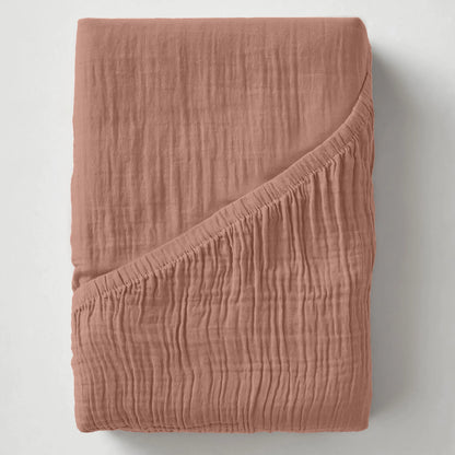 Het Mousseline Hoeslaken - Dusty-Rose, een rozekleurig hoeslaken van mousseline katoen ademend beddengoed, wordt getoond op een wit oppervlak met een hoek naar achteren gevouwen om de dikte en stofdetails te laten zien.