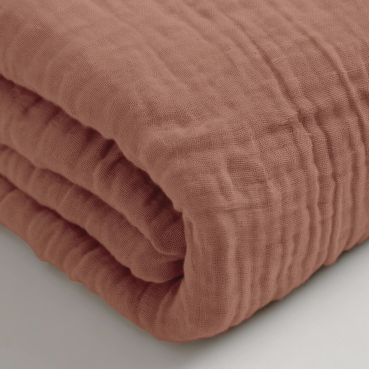 Een close-up van de Mousseline Hoeslaken - Dusty-Rose, een gevouwen katoenen laken met structuur op een wit oppervlak -ideaal ademend beddengoed voor sereen comfort.