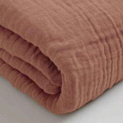 Een close-up van de Mousseline Hoeslaken - Dusty-Rose, een gevouwen katoenen laken met structuur op een wit oppervlak -ideaal ademend beddengoed voor sereen comfort.
