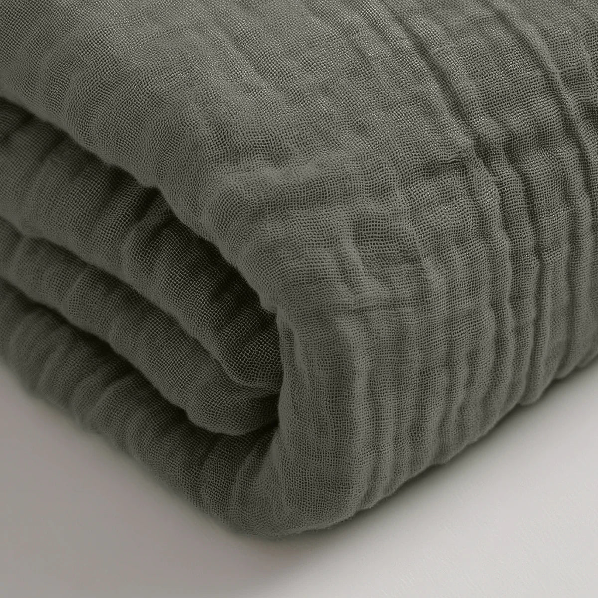 Een gevouwen, grijsgroene Mousseline Hoeslaken staat op een wit oppervlak, waardoor het ideaal is als beddengoed voor elke Japans geïnspireerde ruimte.