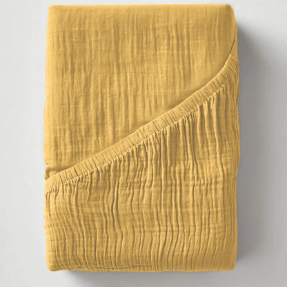 De Mousseline Hoeslaken in okergeel, met zijn omgeslagen hoek die zachte, ademende lagen onthult, maakt sereen comfort gemakkelijk - perfect als ademend bedgoed.