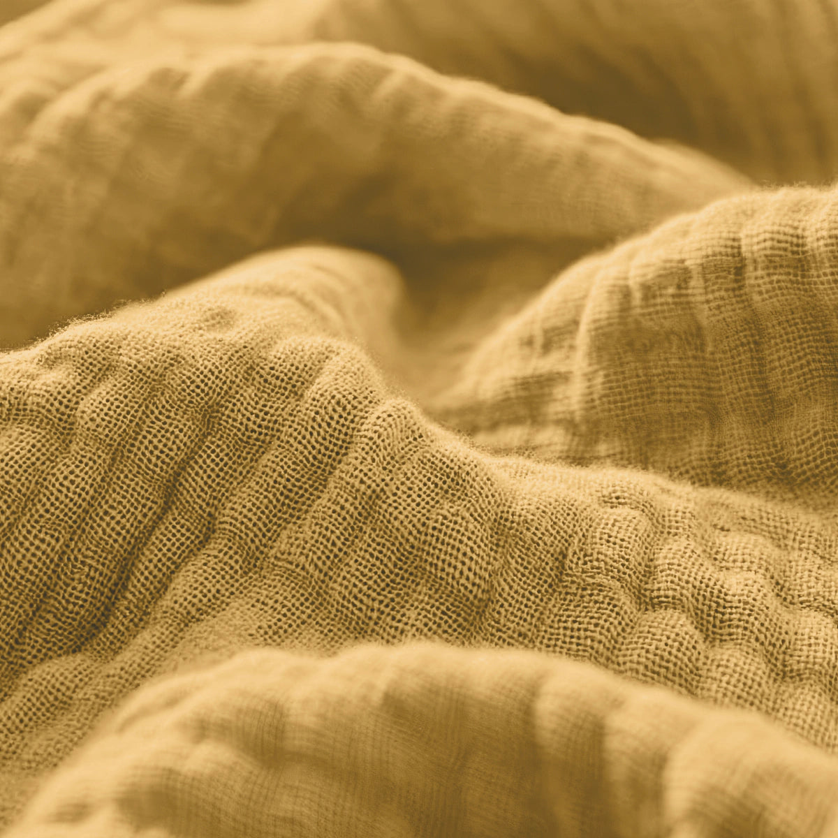 Close-up van de okergele Mousseline Hoeslaken die de textuur, het ademende katoen met de zachte plooien en het zichtbare weefsel laat zien - ideaal voor een Japandi-stijl of luchtig beddengoed.