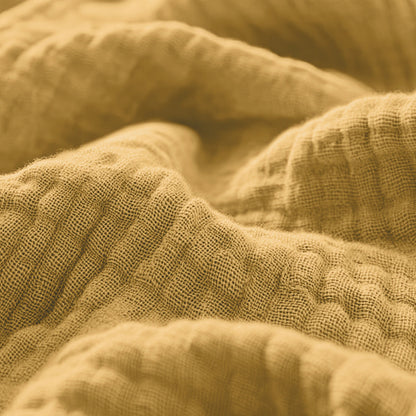 Close-up van de okergele Mousseline Hoeslaken die de textuur, het ademende katoen met de zachte plooien en het zichtbare weefsel laat zien - ideaal voor een Japandi-stijl of luchtig beddengoed.