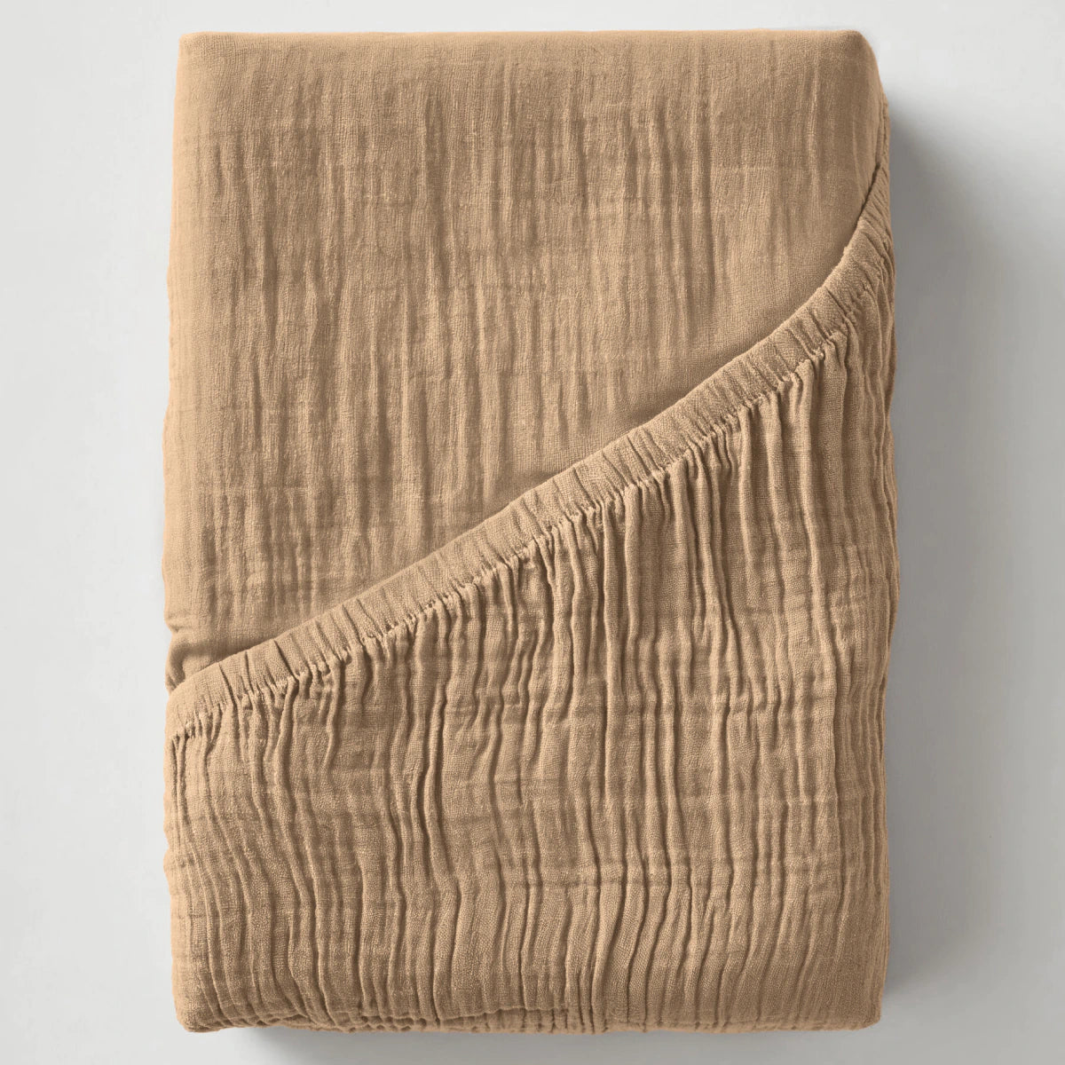 Een gevouwen Mousseline Hoeslaken - Taupe, gestructureerd en lichtbruin met een hoek omgeslagen, wordt weergegeven op een effen, lichte achtergrond.