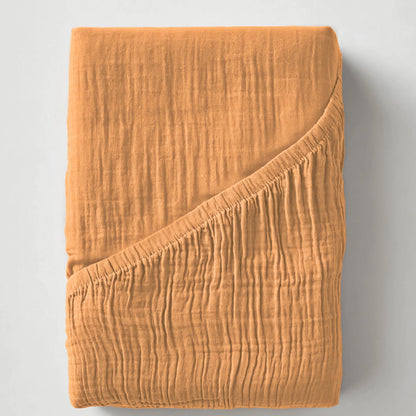 Het Mousseline Hoeslaken - Terra is een gevouwen, gestructureerd oranje mousseline katoenen laken met een omgeslagen hoek op een neutrale achtergrond - ideaal voor liefhebbers van de Japanse stijl die op zoek zijn naar ademend beddengoed.