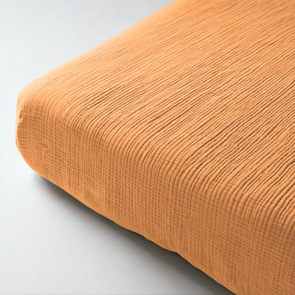 Close-up van een netjes opgemaakt bed met de Mousseline Hoeslaken - Terra, een oranje geribd hoeslaken van zacht mousseline katoen, op een lichte ondergrond voor een uitnodigende uitstraling.