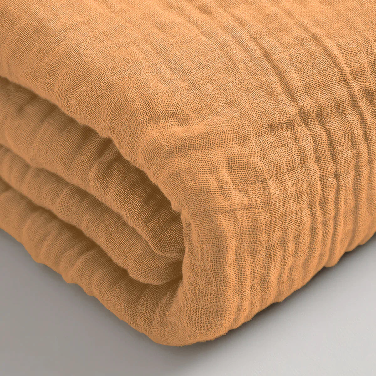 Een close-up van het Mousseline Hoeslaken - Terra, een gevouwen hoeslaken van lichtbruin katoen met textuur op een licht oppervlak, dat de ademende stof benadrukt - perfect voor serene slaapkamers in Japanse stijl.