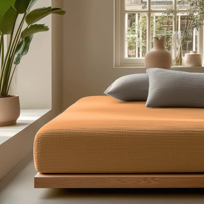 Minimalistische slaapkamer met een laag houten bedframe, oranje sprei, Mousseline Hoeslaken - Terra, twee grijze kussens, potplanten en keramische vazen bij een raam dat natuurlijk licht binnenlaat voor een ademend bedgoed.