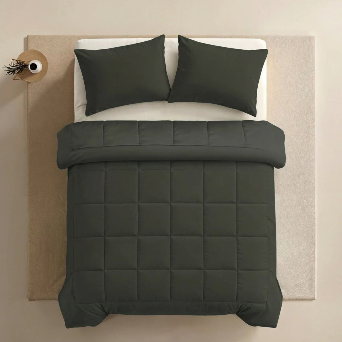 Een keurig opgemaakt bed met donkergroen superzacht katoen beddengoed en twee Zelesta Pure Uni kussenslopen (Olijfzwart) op een licht vloerkleed, van bovenaf gezien. Een klein bijzettafeltje met een vaas en plant staat naast het bed.