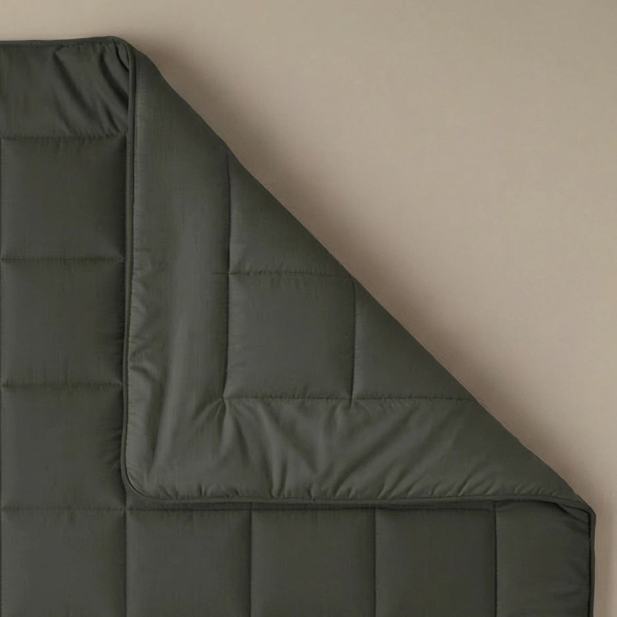 Het Zelesta Pure Uni Dekbed in olijfzwart is netjes uitgespreid op een beige oppervlak met een omgeslagen hoek. De gewatteerde textuur en stiksels zorgen voor een ademend slaapcomfort.