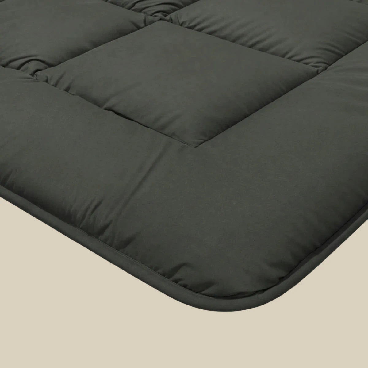 Close-up van een donkergrijze gewatteerde matrastopper met vierkante stiksels, getoond op een beige ondergrond - ideaal voor ultiem slaapcomfort en perfect gecombineerd met het Zelesta Pure Uni Dekbed - Olijfzwart.