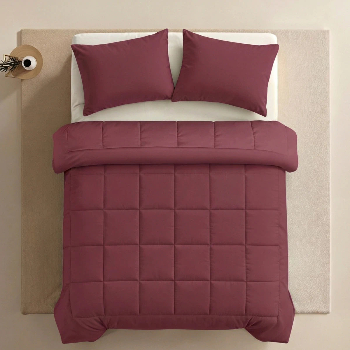 Een netjes opgemaakt bed met het Zelesta Pure Uni Dekbed in Bordeaux Rood en bijpassende kussens, op een crèmekleurig vloerkleed met een klein rond nachtkastje ernaast, biedt ademend slaapcomfort met duurzame dekbedden.
