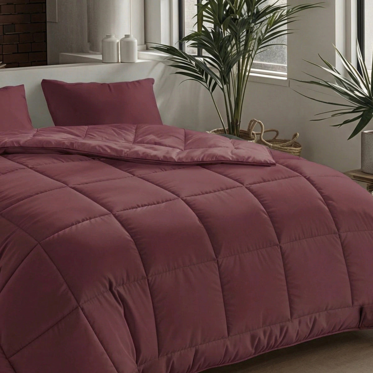 Een netjes opgemaakt bed met het Zelesta Pure Uni Dekbed in Bordeaux Rood en bijpassende kussens biedt een adembenemend slaapcomfort, bij een raam met potplanten en decoratieve items op een bijzettafel.