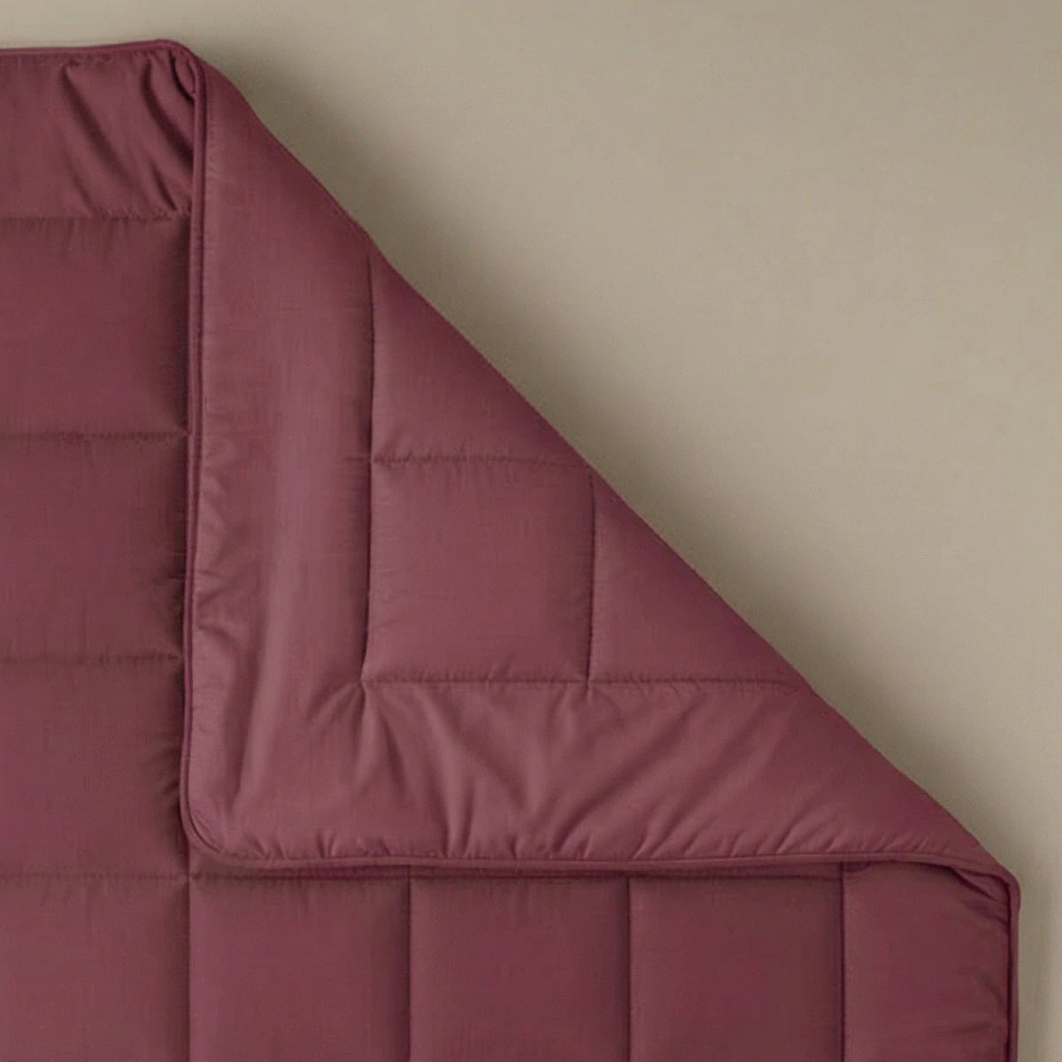 Een Zelesta Pure Uni Dekbed in bordeaux rood is plat neergelegd op een beige ondergrond, met een hoek omgevouwen om de stiksels en stofstructuur te onthullen voor een ademend slaapcomfort.